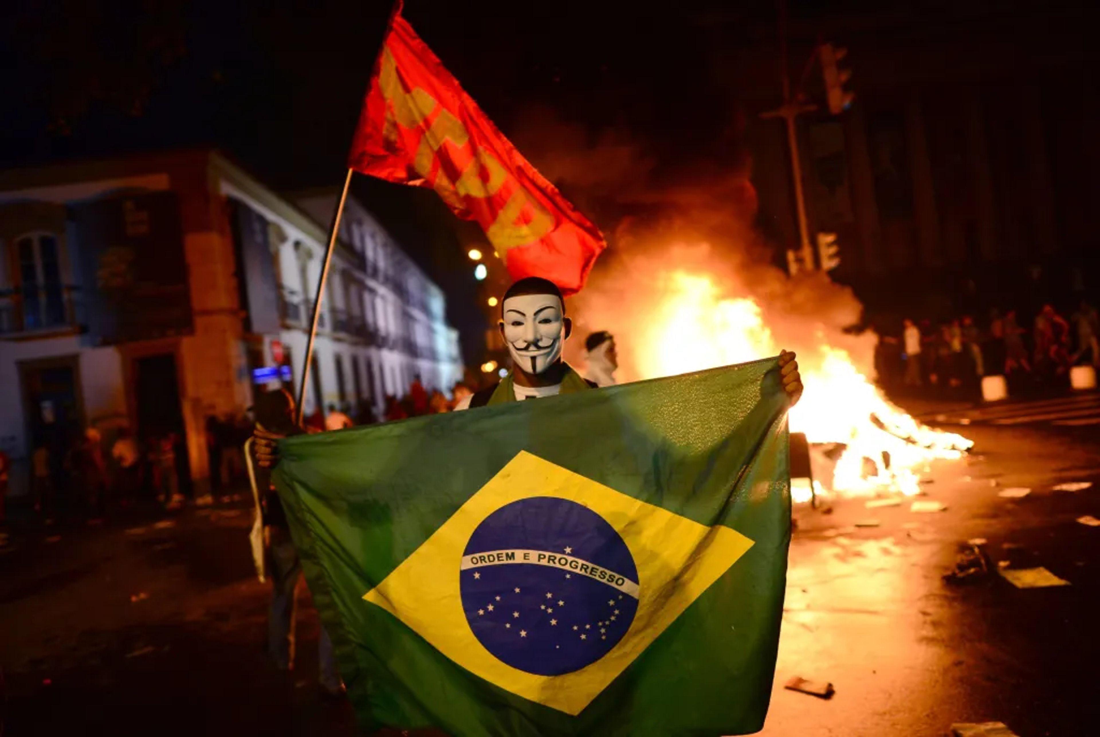 A man stands in front of a fire in the street, holding the Brazillian flag while wearing a 'Guy Fawkes' mask.